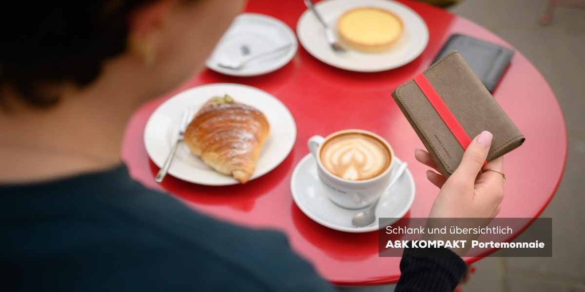 Ein mittelgroßes Portemonnaie in Braun/Rot, platziert auf einem Kaffeetisch mit einer roten Tischdecke. Im Hintergrund befindet sich ein gedeckter Tisch mit einem Stück Kuchen und einer Tasse Kaffee, was die Gemütlichkeit und Vielseitigkeit des Produkts b